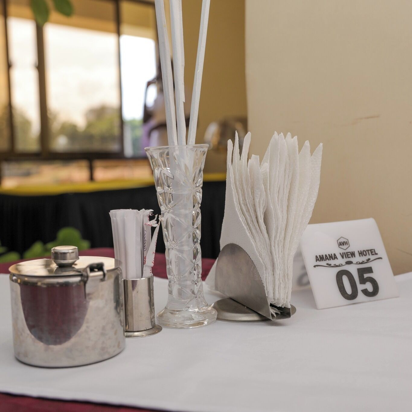 A table with a glass vase, a metal container, and a sign reading "AMANA VIEW HOTEL 05".