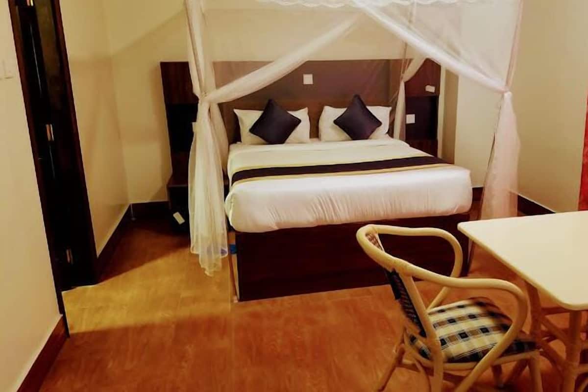 A single bed with a canopy, a small table, and a chair in a room with wooden flooring and a plain wall.