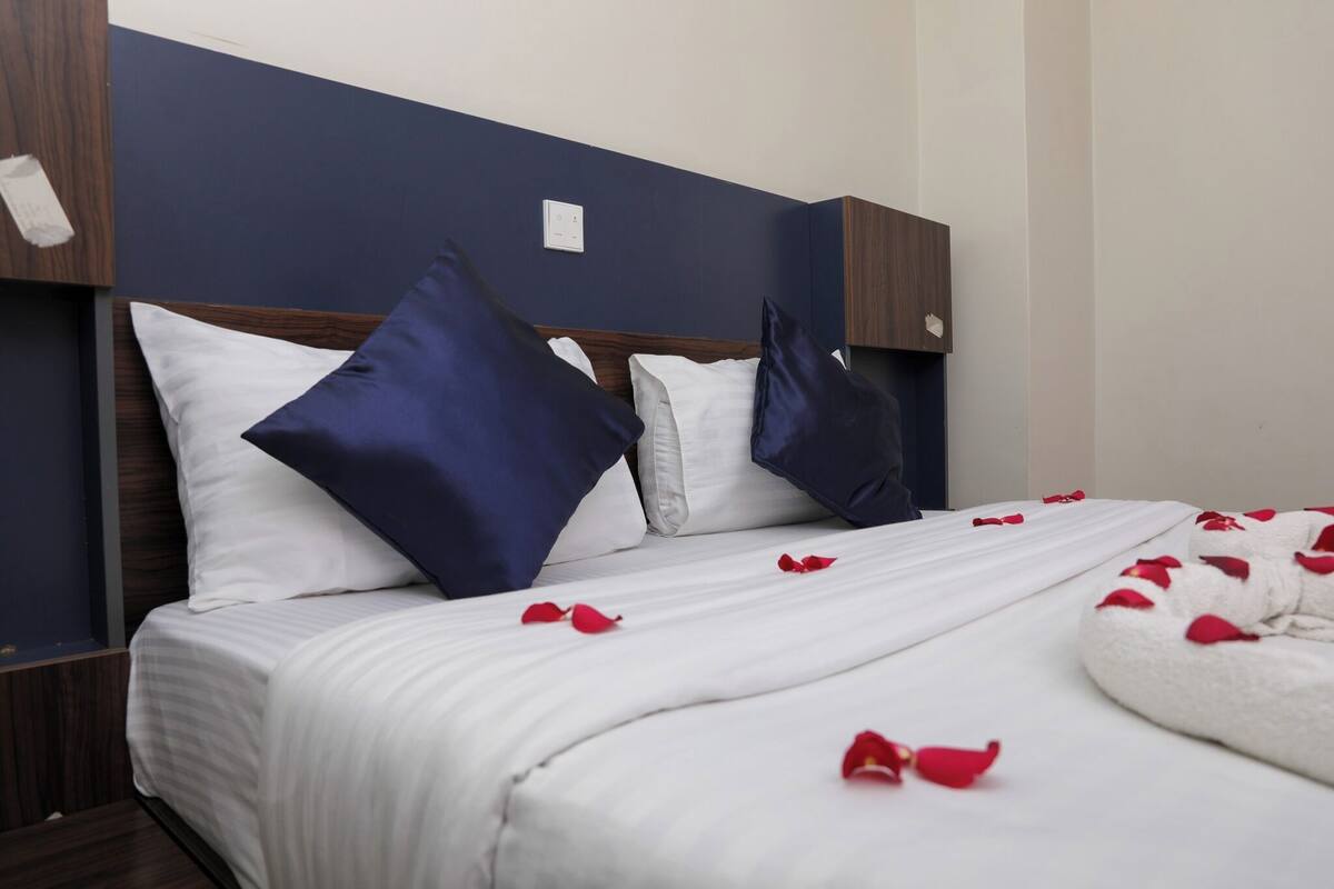 A neatly made bed with white linens and blue pillows, a wooden headboard, and a white throw blanket with red rose petals.