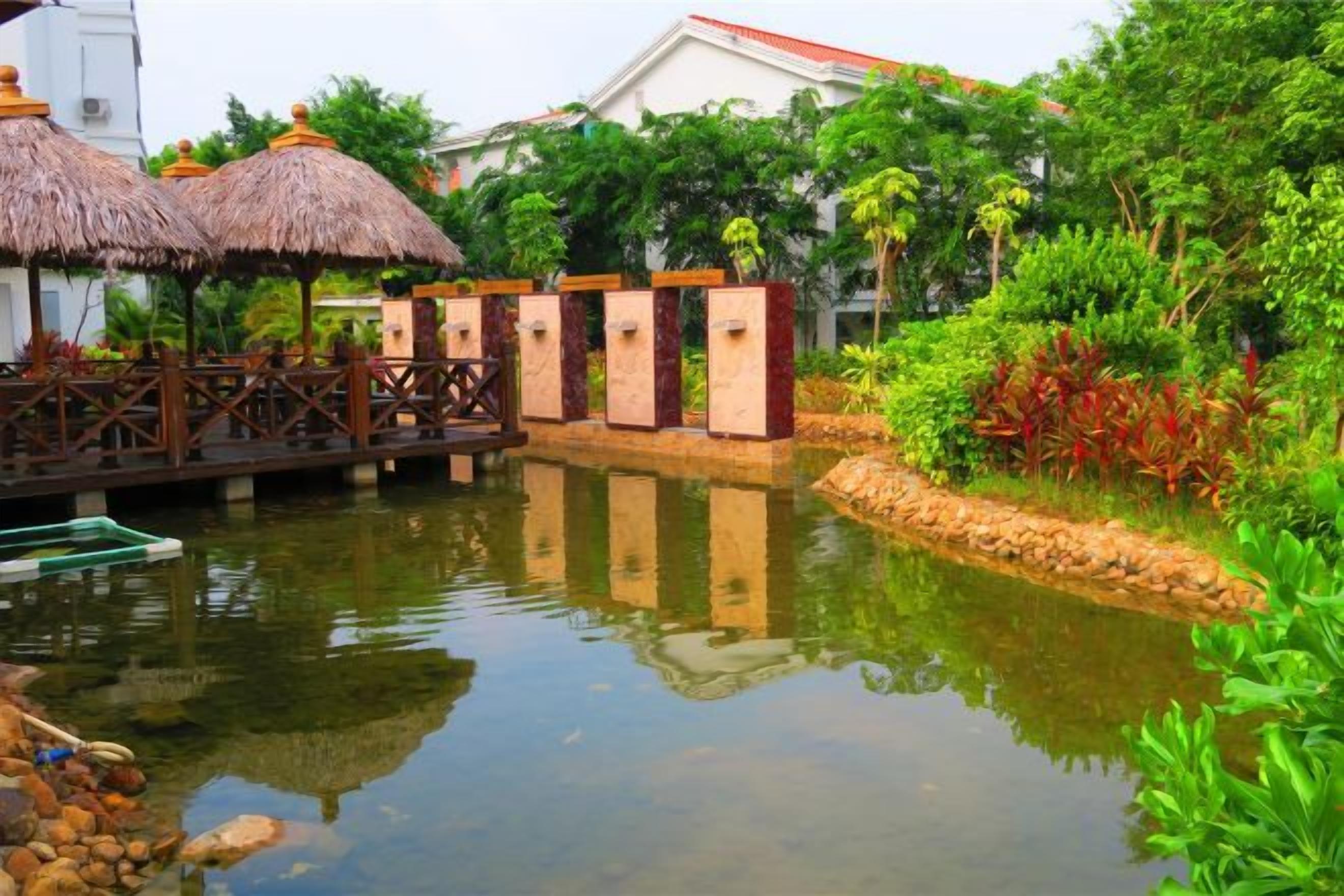 A tranquil pond with a wooden bridge, surrounded by lush greenery and thatched-roof structures.