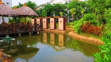 A tranquil pond with a wooden bridge, surrounded by lush greenery and thatched-roof structures.
