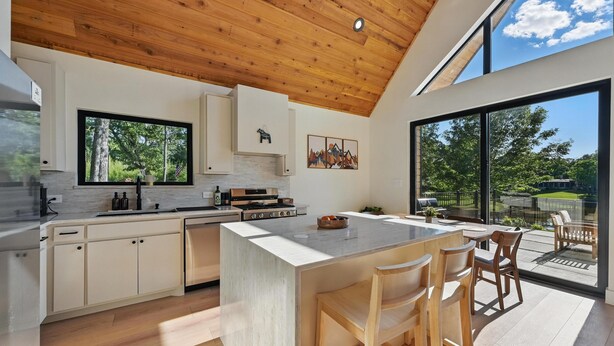 A modern kitchen with a large island, wooden ceiling, and large windows offering a view of the outdoors.