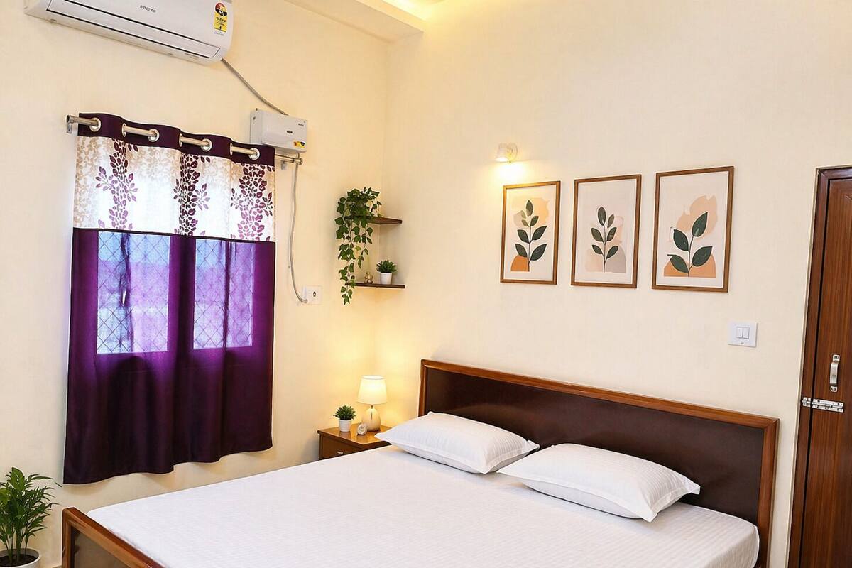 Ein Hotelzimmer mit Bett, Vorhängen am Fenster, einer Nachttischlampe, einer Wand-Klimaanlage und gerahmten Kunstwerken.