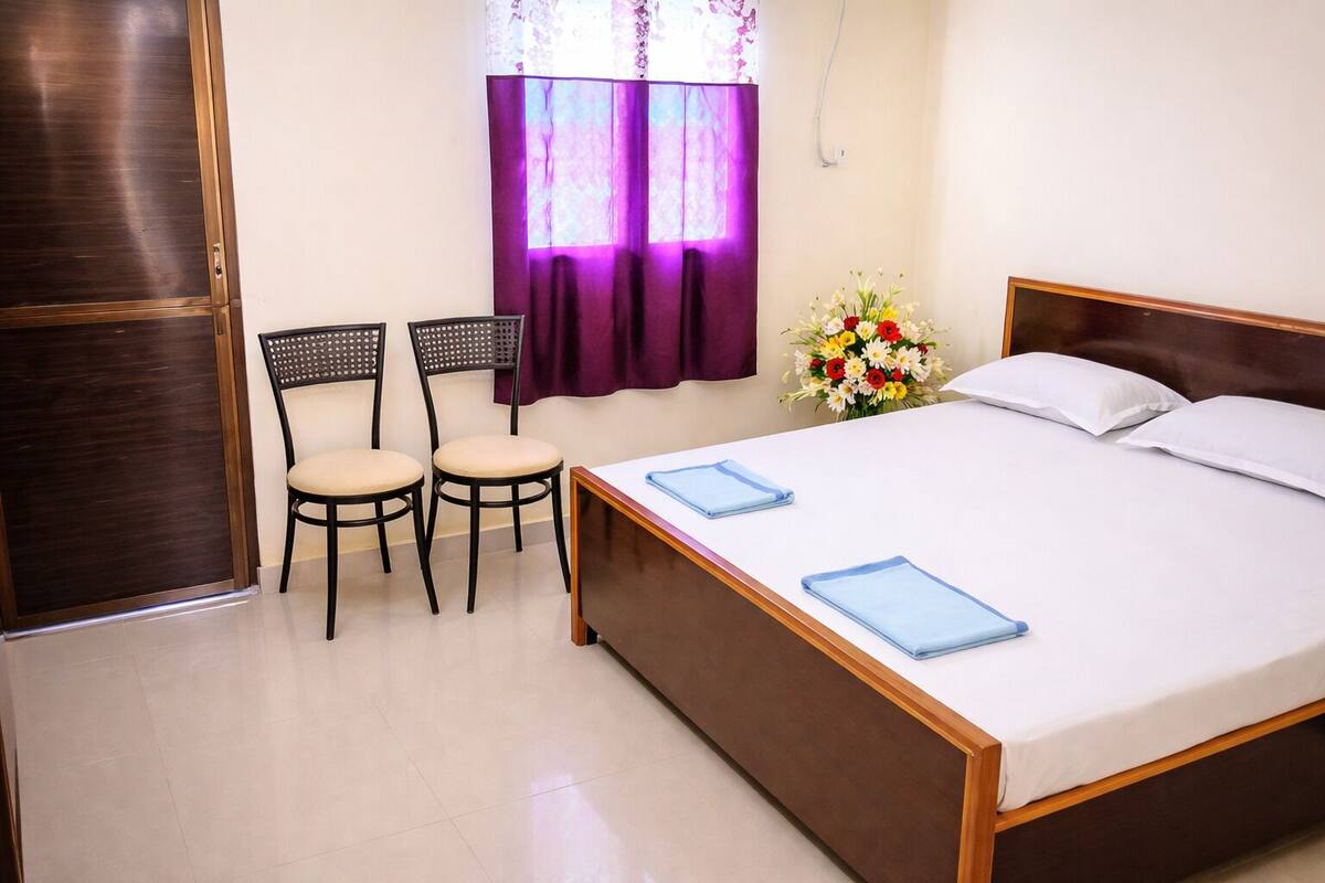 Ein Schlafzimmer mit einem Bett, zwei Stühlen, einem Fenster mit violetten Vorhängen und einer Vase mit Blumen.