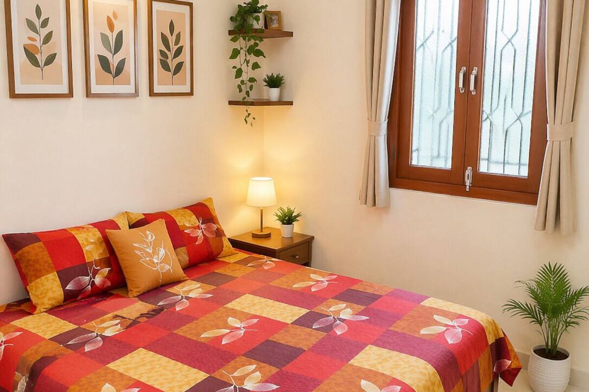 Ein Schlafzimmer mit Bett, Nachttisch mit Lampe, Fenster mit Vorhängen und an der Wand befestigten Regalen mit Pflanzen.