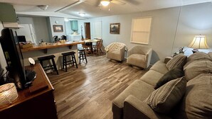 Ein Wohnzimmer mit einer Couch, Sesseln, einem Esstisch und einer Küchenzeile mit Kühlschrank und Arbeitsplatte.