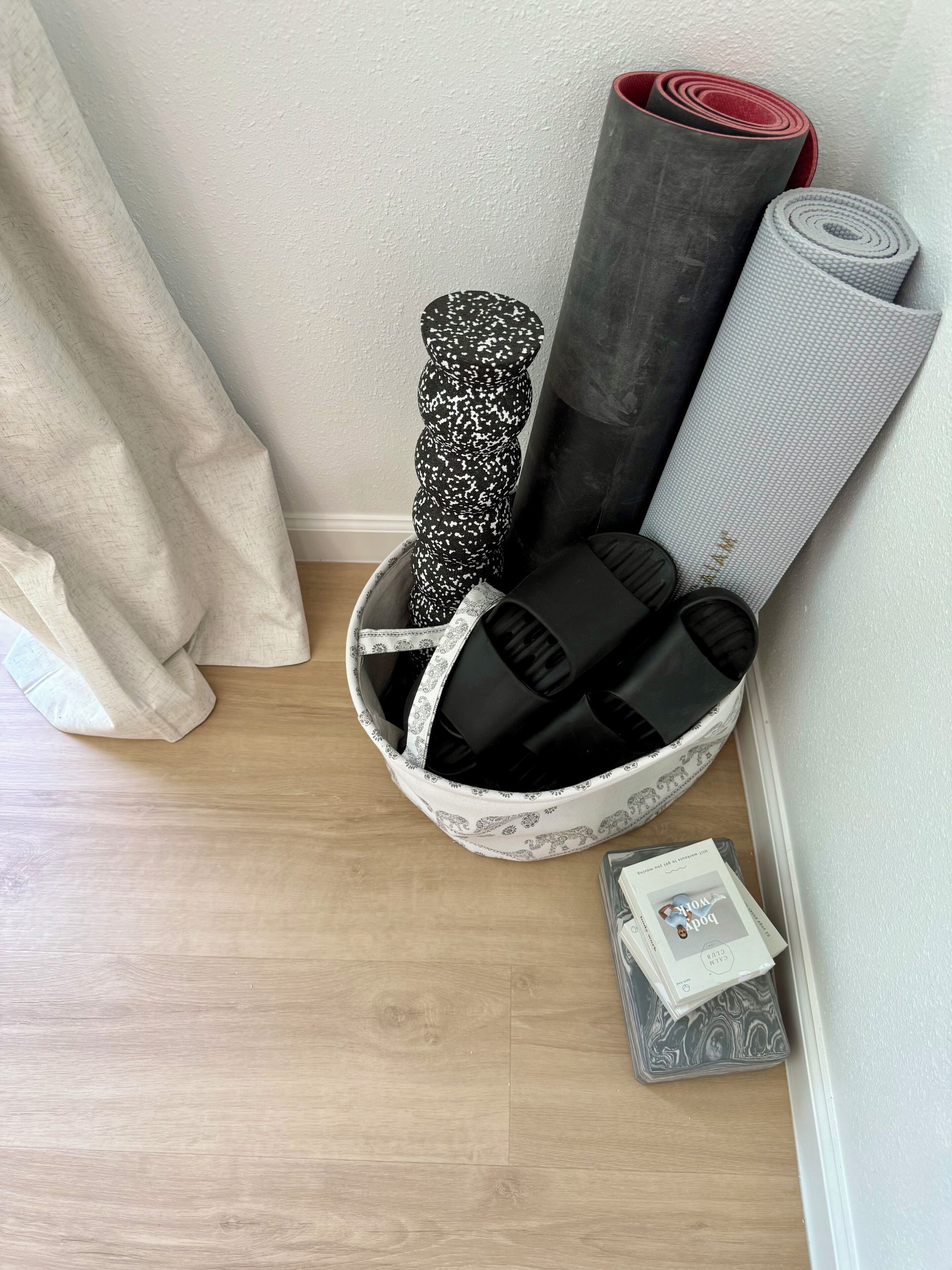Eine Raumecke mit einem weißen Korb, der Yogamatten und ein Buch mit dem Titel „Book of Work“ enthält.