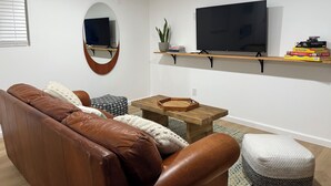 A living room with a brown leather sofa, a round mirror, a wooden coffee table, and a flat-screen TV mounted on a wall. - Cozy casita nestled in Laketree Estates, 5 min walk to Monserate Winery. (Fallbrook)