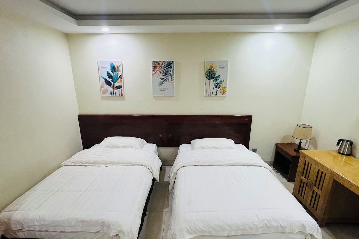 A hotel room with two single beds, a wooden headboard, a small desk with a lamp, and three wall-mounted artworks.