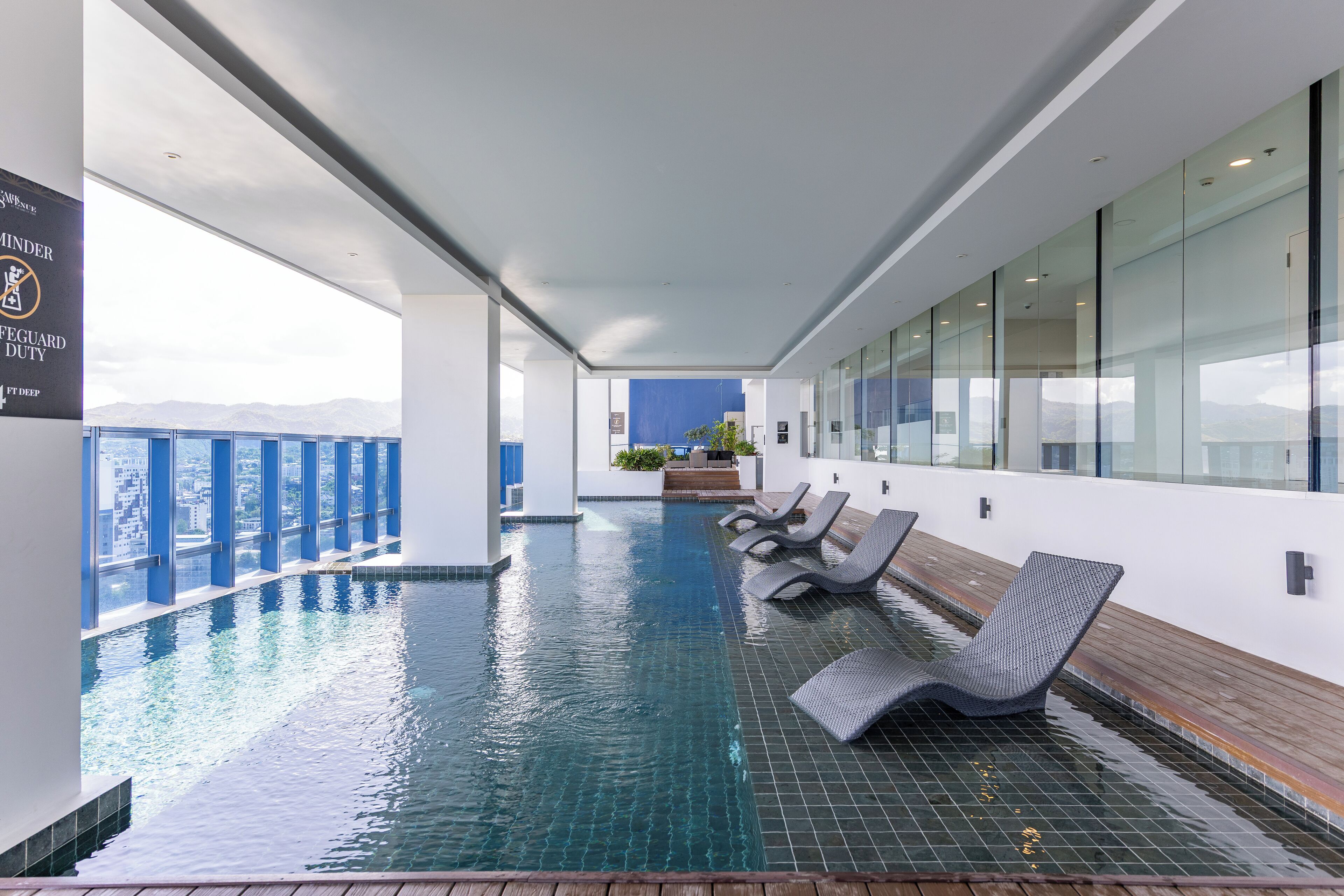 Una piscina interior moderna con paredes de vidrio transparente, suelo de baldosas y varias sillas de descanso.
