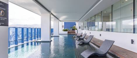 Una piscina interior moderna con paredes de vidrio transparente, suelo de baldosas y varias sillas de descanso.