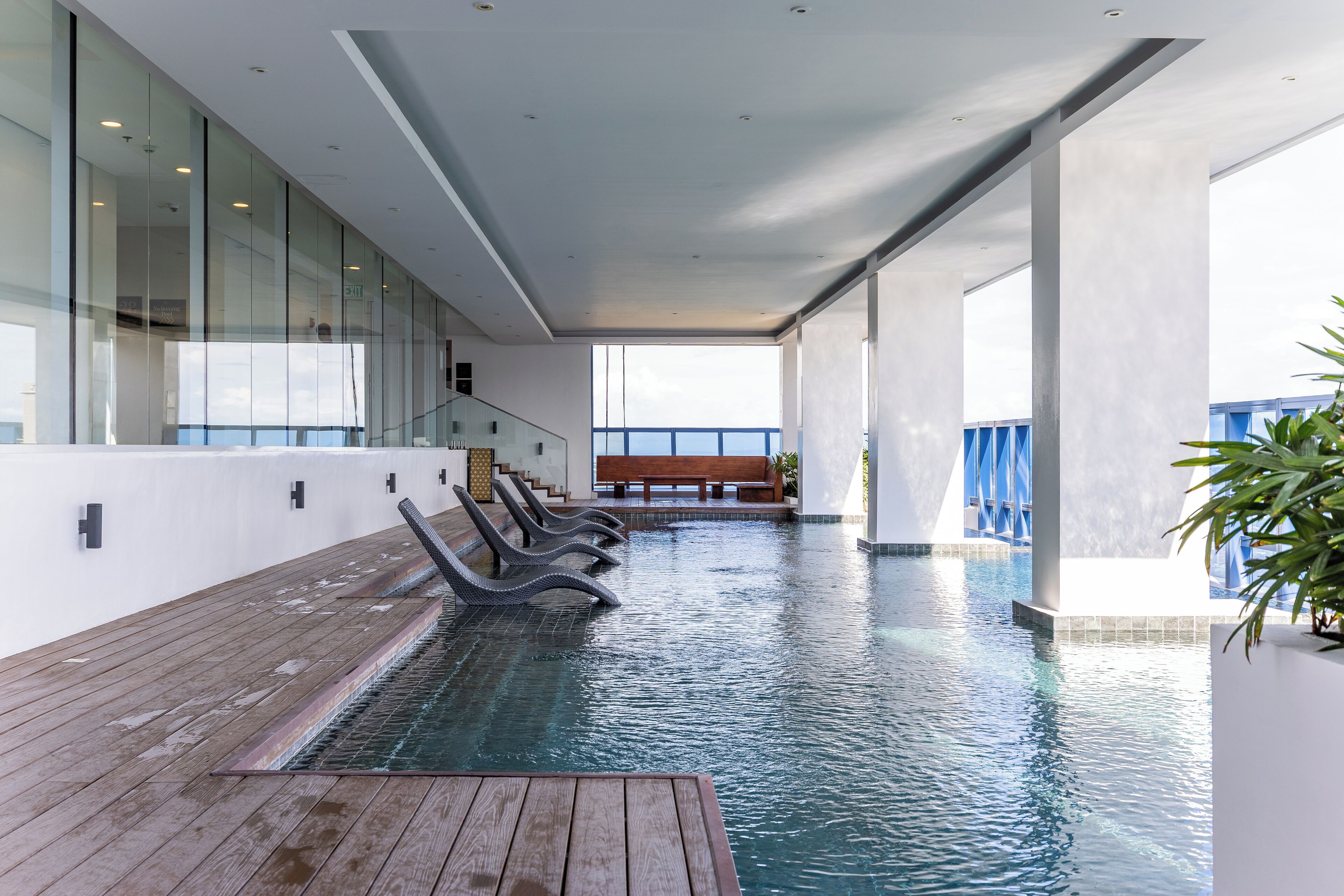 A modern indoor pool area with clear glass walls, a wooden deck, and a bench.