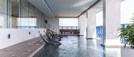 A modern indoor pool area with clear glass walls, a wooden deck, and a bench.