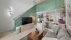 Una sala de estar moderna con televisor, sofá y mesa de centro. Una escalera conduce a un nivel superior con cocina y comedor.