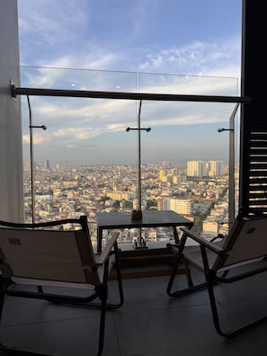 A balcony with two chairs and a table, overlooking a cityscape with buildings and a clear sky. - X-Stay 2908 Lotus (Hai Phong)