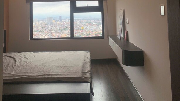 Un dormitorio con cama, ventana con vista a la ciudad, lámpara de techo y estante fijado a la pared.