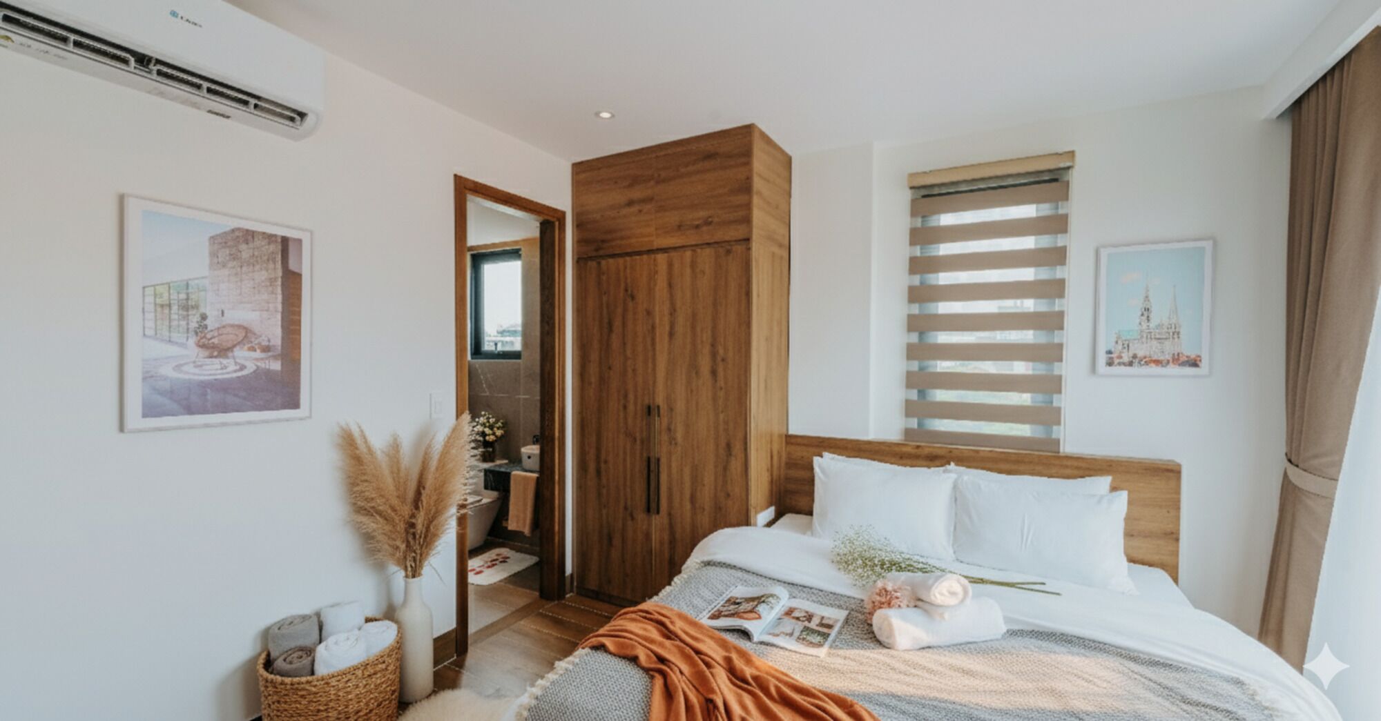Une chambre à coucher moderne avec une tête de lit en bois, une grande fenêtre avec des stores, une table de chevet ornée d’un vase contenant des plumes d’herbe des pampas et un lit recouvert de linge de maison blanc.