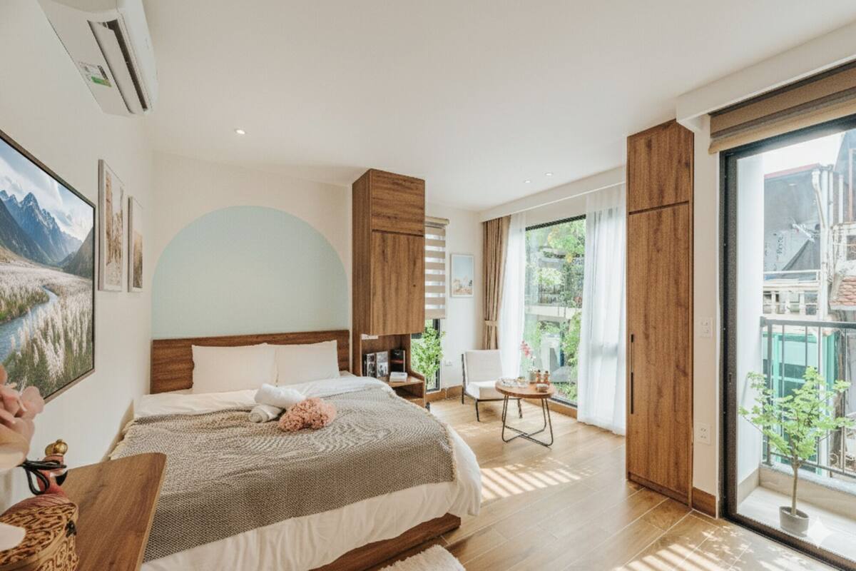 A modern bedroom with a large bed, a wooden wardrobe, a small table, and a balcony with a view of greenery.