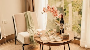 Um quarto acolhedor com piso de madeira, uma mesa redonda com uma revista, uma garrafa de vinho e um vaso de flores cor-de-rosa.