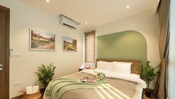 Quarto com cama, dois aparelhos de ar condicionado fixados na parede, janela com persianas e duas imagens emolduradas de paisagens.