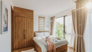 A bedroom with a wooden wardrobe, a bed with a beige blanket, and a large window with curtains.