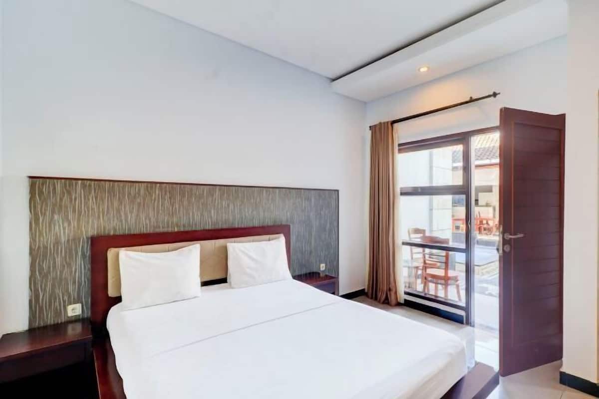 A hotel room with a bed, a nightstand, a window with curtains, and a door leading to an outdoor area.
