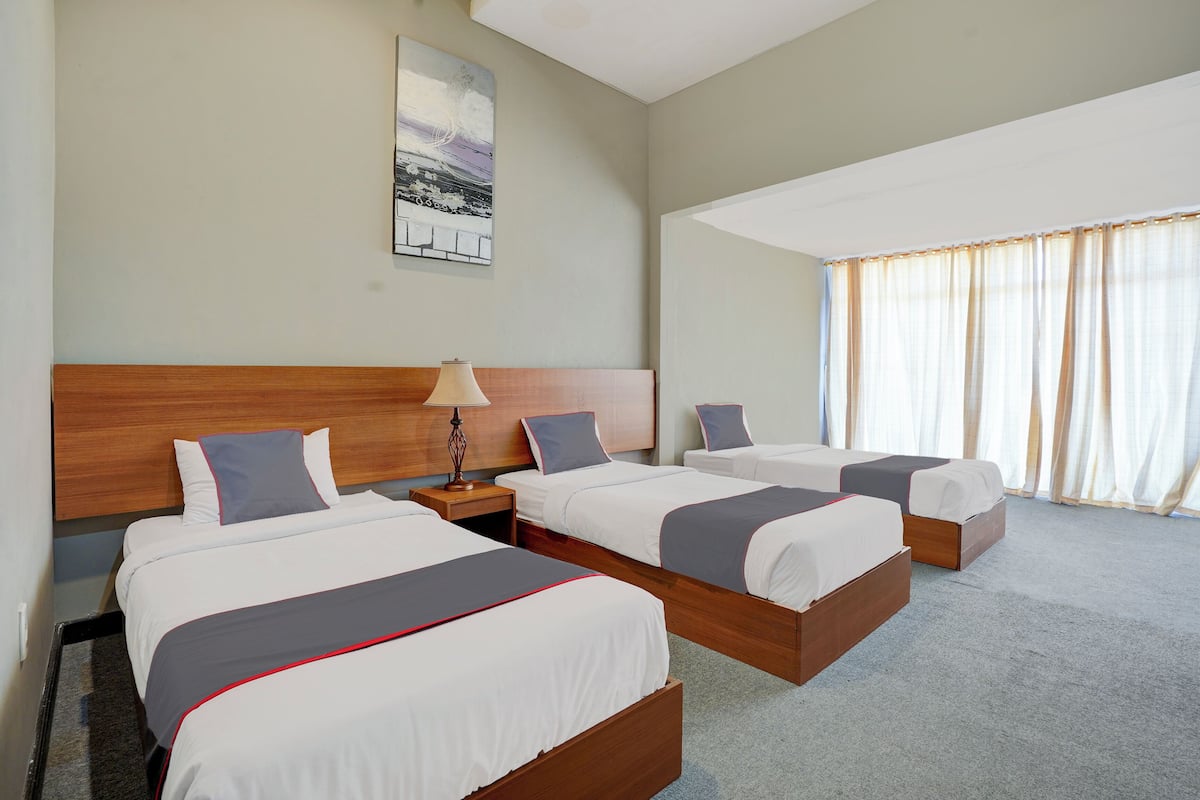A hotel room with three beds, a wooden headboard, a nightstand with a lamp, and a large window with curtains.