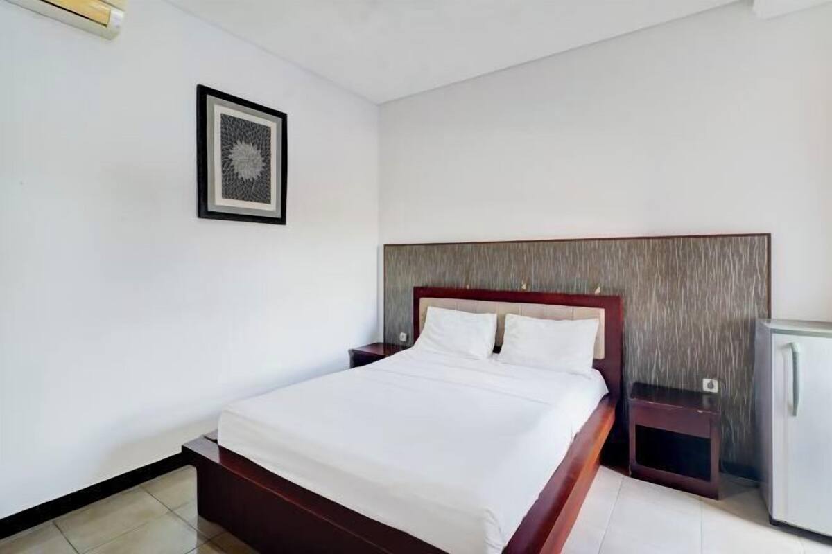 A hotel room with a bed, a nightstand, a small table, and a refrigerator.
