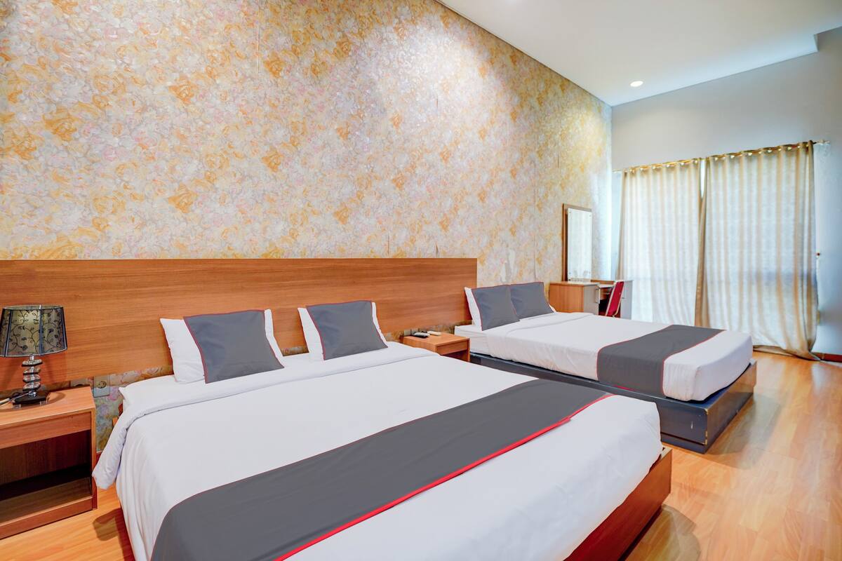 A hotel room with two beds, a wooden headboard, a nightstand with a lamp, and a large window with curtains.