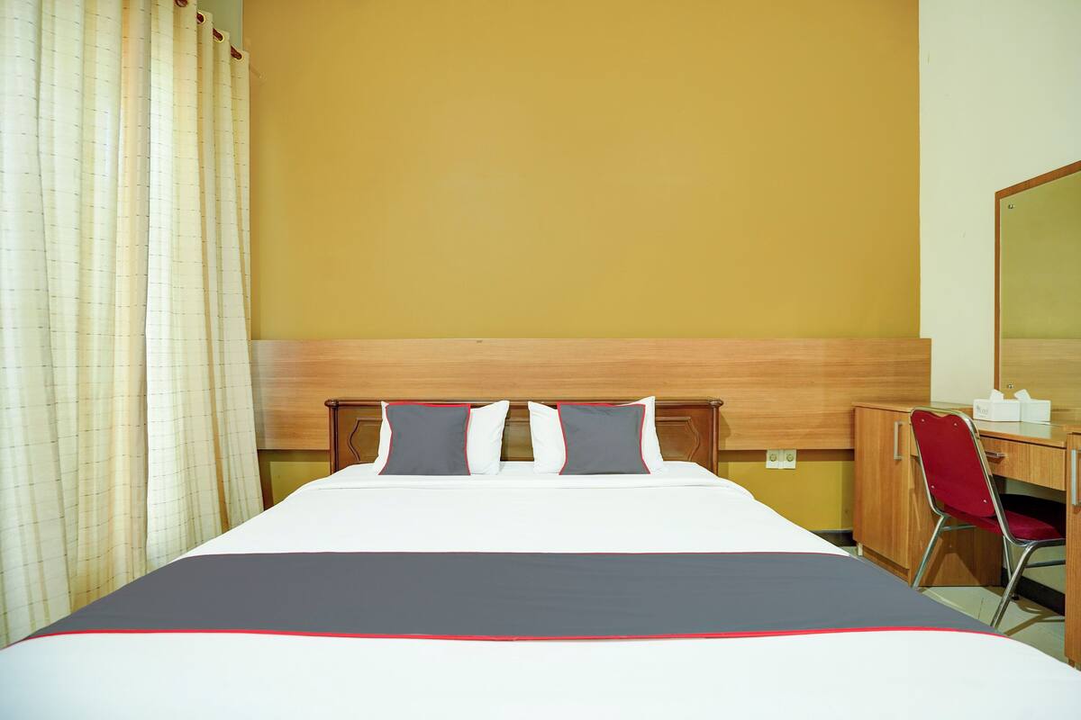 A hotel room with a bed, a desk, a chair, a mirror, and a curtain.