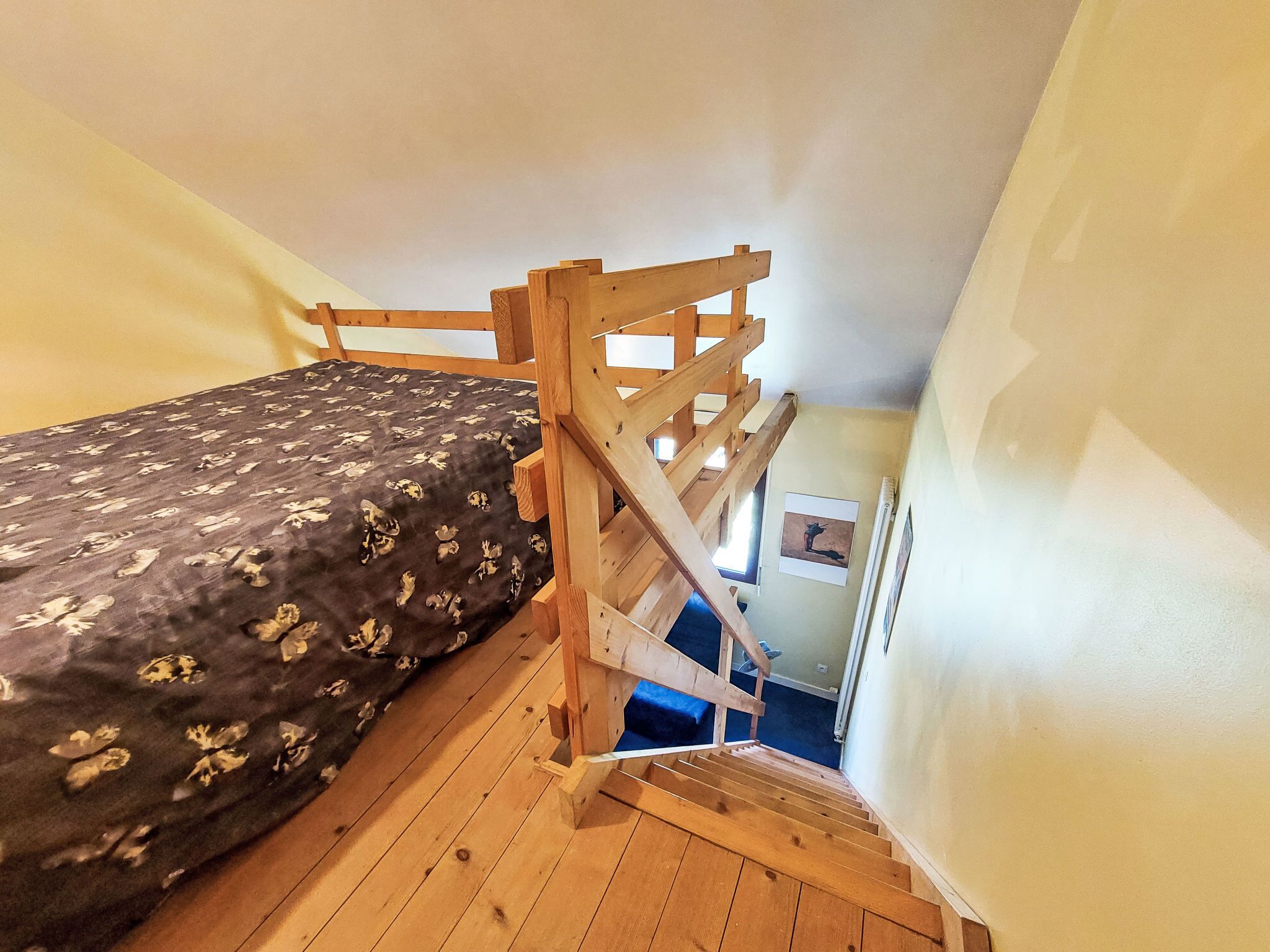 A small, single-bed room with a wooden bunk bed structure, a patterned bedspread, and a window with a picture on the wall.