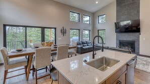 Una cocina moderna con una isla grande, un comedor con mesa y sillas, una chimenea y ventanas amplias que ofrecen vistas al exterior.