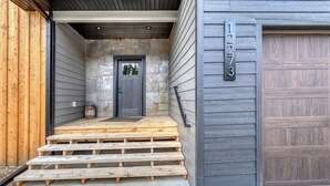 Entrada de una casa con un muro de piedra, una puerta gris y una escalera de madera que lleva a un garaje.