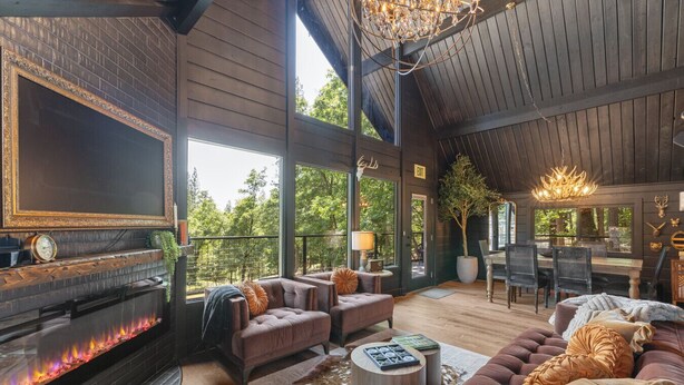 A spacious living area with a fireplace, leather sofas, and a dining area with a chandelier. - Custom Mountain Home · Hot Tub · Waterfall · 2.5 Acres · EV Charger (Placerville)