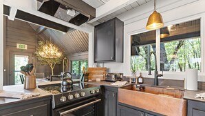 A modern kitchen with a gas stove, oven, and copper sink. There is a wooden dining table with chairs, a chandelier, and a view of trees through large windows. - Custom Mountain Home · Hot Tub · Waterfall · 2.5 Acres · EV Charger (Placerville)