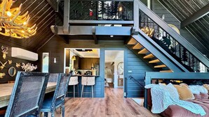 A spacious interior with a wooden floor, a dining area with chairs and a table, a bed with pillows, and a staircase leading to an upper level. - Custom Mountain Home · Hot Tub · Waterfall · 2.5 Acres · EV Charger (Placerville)