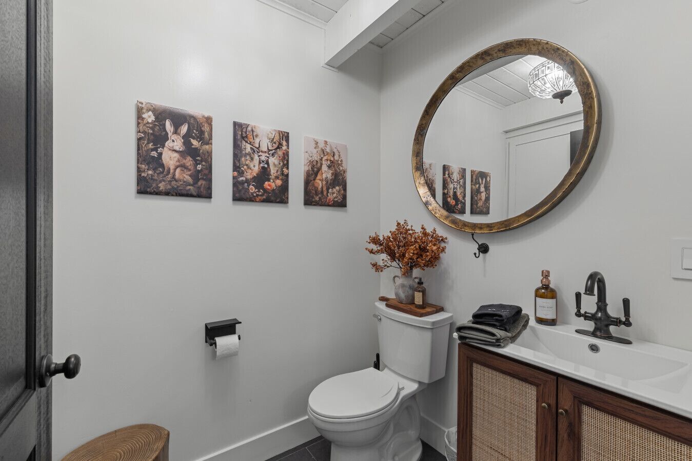A bathroom with a toilet, sink, and mirror. There are three wall-mounted paintings above the toilet.