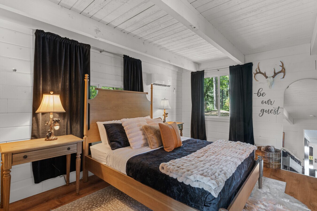 A bedroom with a wooden bed, dark curtains, a nightstand with a lamp, and a wall with a 'be our guest' sign.