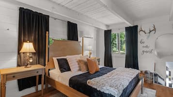 A bedroom with a wooden bed, dark curtains, a nightstand with a lamp, and a wall with a 'be our guest' sign.