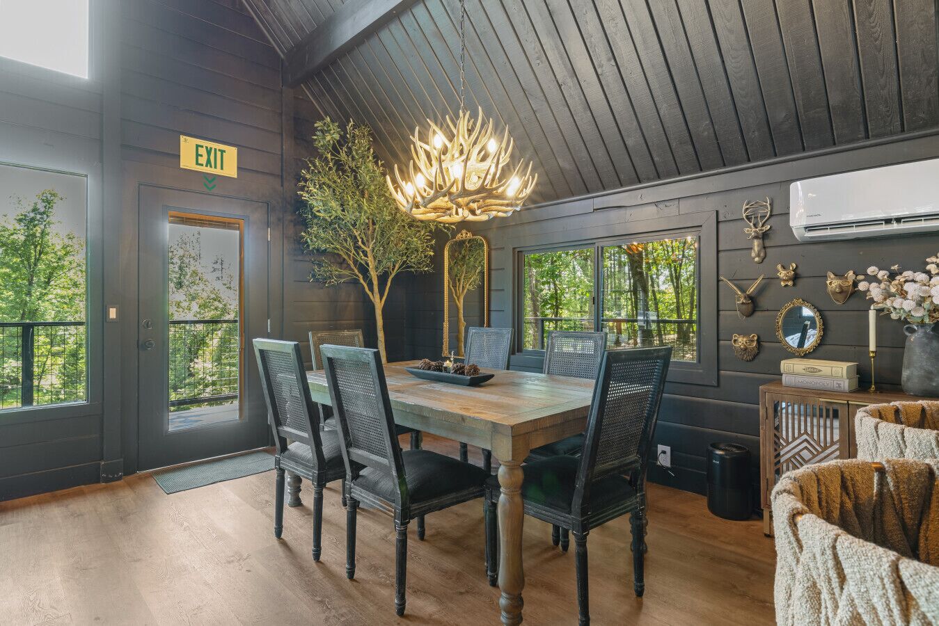 A dining area with a wooden table, four chairs, a chandelier, and a view of trees through large windows.