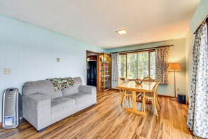 Habitación con sofá gris, mesa de comedor de madera y sillas. Se observa un calefactor, una ventana con cortinas y una puerta que da a otra habitación.