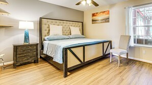 Quarto com cama, mesa de cabeceira, abajur, cadeira, ventilador de teto, janela e obra de arte.