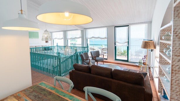 A spacious living area with a view of the sea, a balcony with teal railings, and a dining area with a wooden table and chairs.