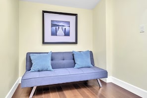 Quarto com sofá cinzento, duas almofadas azuis, um quadro na parede e chão de madeira.