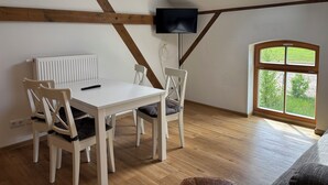 A room with a sloped ceiling, a white table with four chairs, a TV mounted on the wall, and a large window with a view of greenery. - Cozy vacation apartment / fitter's room for 1-6 persons near Berlin FeWo 8. (Liebenwalde)