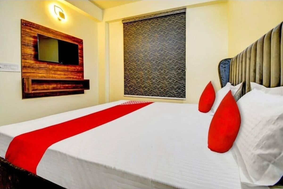 Habitación de hotel con cama, televisor montado, ventana con persiana decorativa y detalle rojo en la cama.