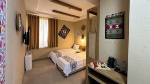 A hotel room with a bed, a bedside table with a kettle and a bottle, a wall-mounted TV, a window with curtains, and a wall hanging. - Hotel Byond Khiva (Khiva)