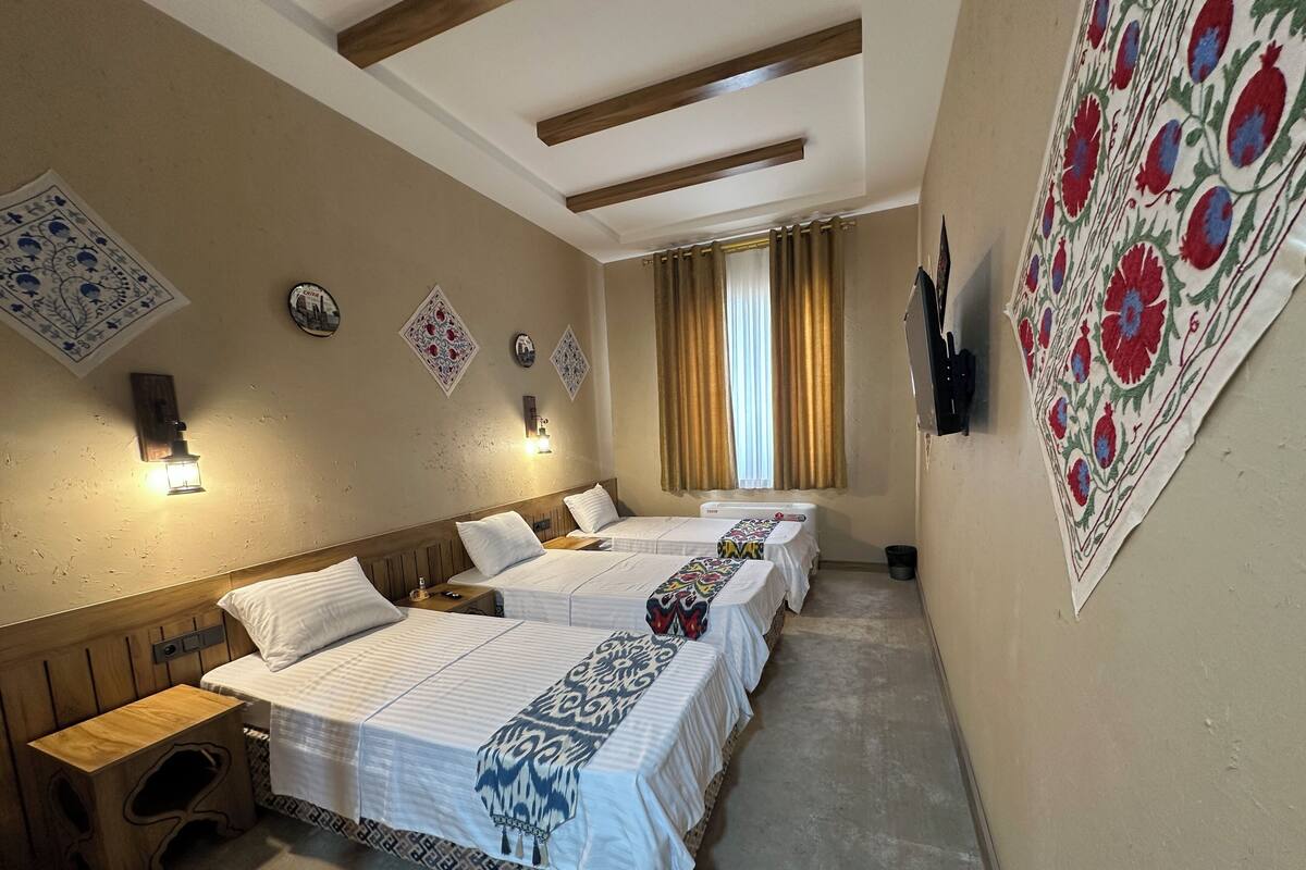 A hotel room with three beds, wooden headboards, a small wooden stool, a wall-mounted TV, and decorative wall art.