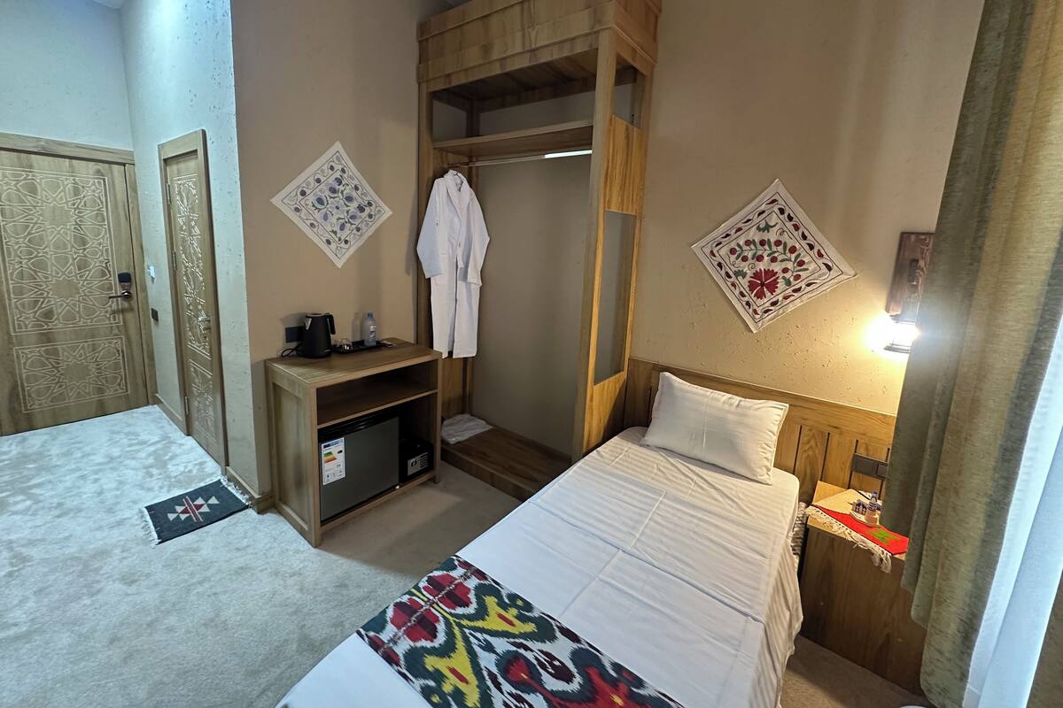 A hotel room with a bed, a wooden wardrobe, a desk with a kettle, and a robe hanging on the wardrobe.