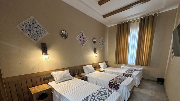 A hotel room with three beds, a wooden headboard, a small wooden bench, and a window with curtains. - Hotel Byond Khiva (Khiva)